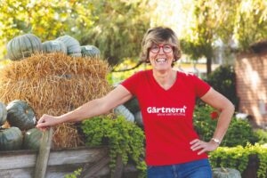 Katrin Iskam lächelt im Garten, trägt ein rotes T-Shirt mit "Gärtnern ohne viel Geschiss" vor Heuballen und Kürbissen