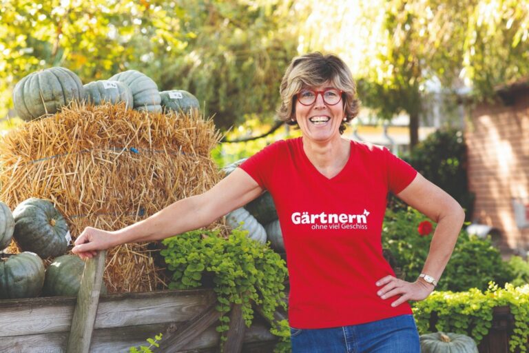 Katrin Iskam lächelt im Garten, trägt ein rotes T-Shirt mit "Gärtnern ohne viel Geschiss" vor Heuballen und Kürbissen
