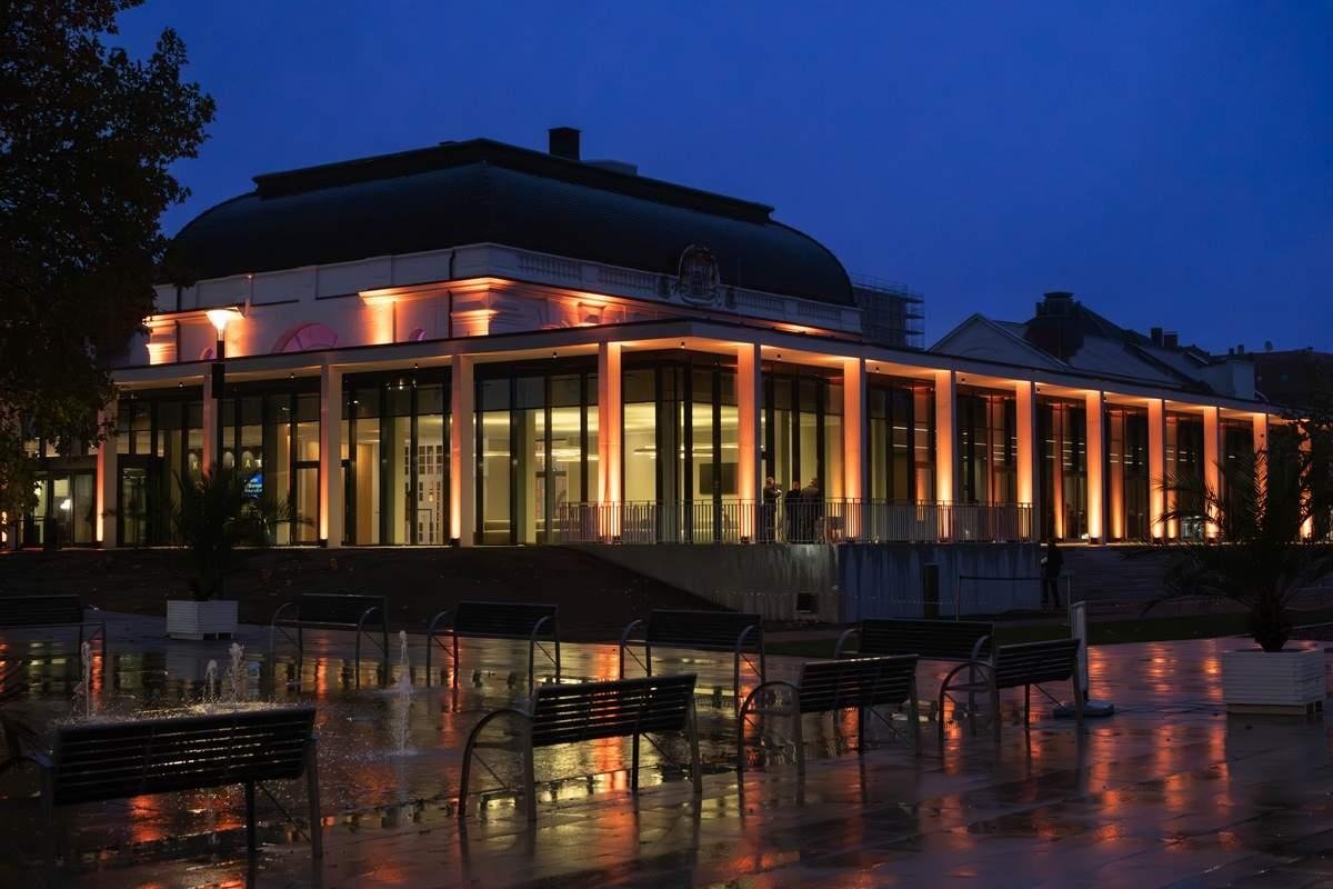 Orange beleuchtetes Gebäude mit Glasfassade und Säulen bei Nacht, davor Platz mit Bänken, Wasserspielen und Personen