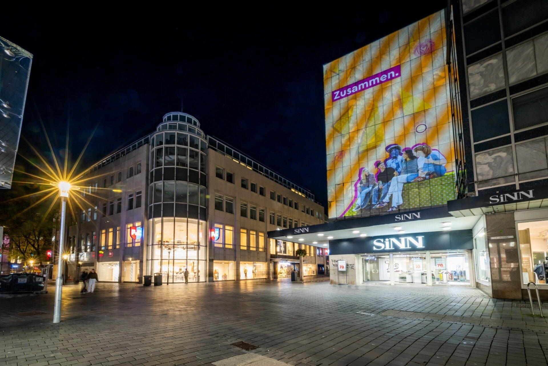 Nächtliche Einkaufsstraße mit Sinn-Gebäude, Menschen und großer "Zusammen." Projektion auf der ehemaligen