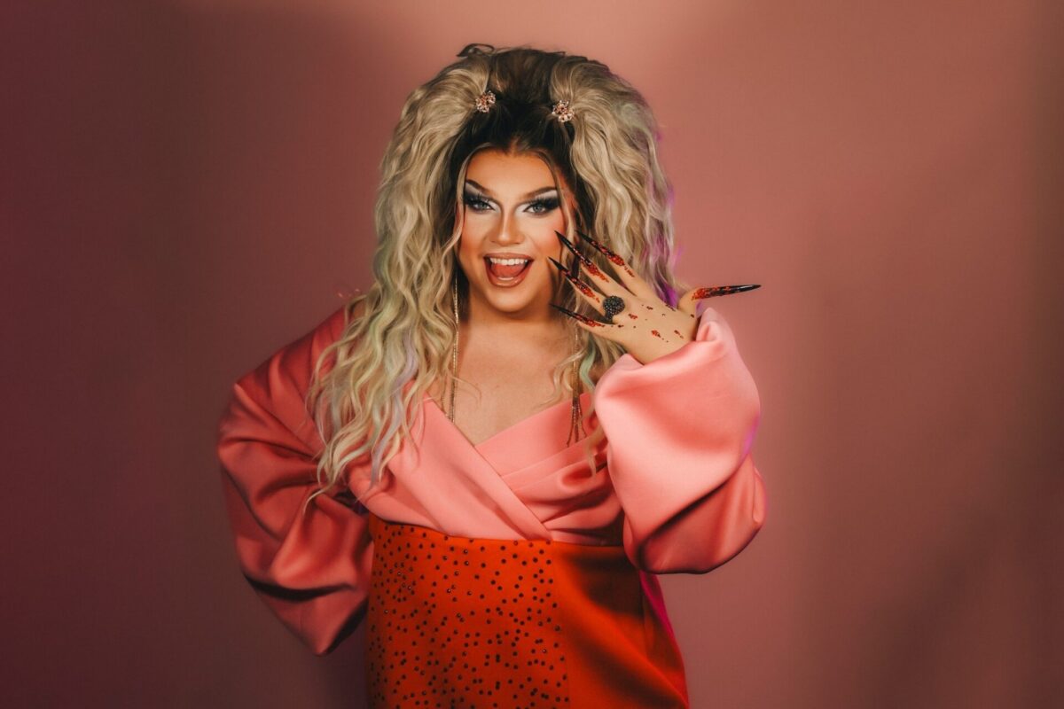 Pam Pengco, eine Drag Queen, mit langer blonder Perücke, pink-orangenem Outfit und glitzernden Krallennägeln lächelt