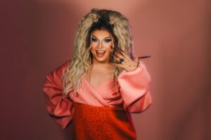 Pam Pengco, eine Drag Queen, mit langer blonder Perücke, pink-orangenem Outfit und glitzernden Krallennägeln lächelt