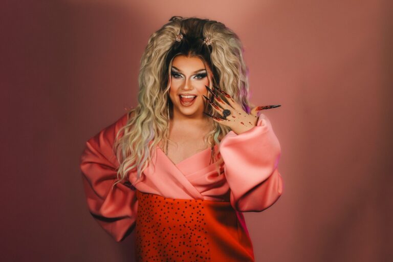 Pam Pengco, eine Drag Queen, mit langer blonder Perücke, pink-orangenem Outfit und glitzernden Krallennägeln lächelt