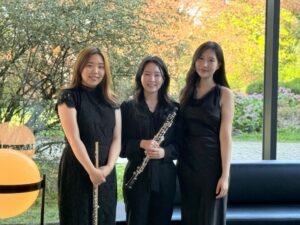 Detmolder Trio: Drei lächelnde Musikerinnen in schwarzer Kleidung mit Flöte und Oboe vor grüner Naturkulisse
