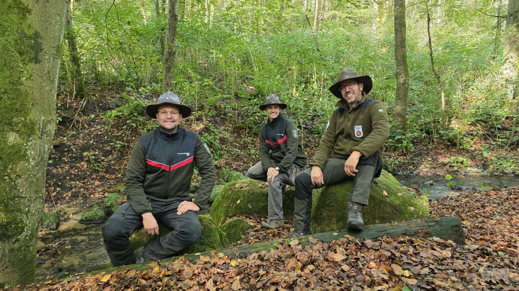 Drei lächelnde Ranger in Uniform sitzen im Herbstwald an einem Bach auf moosbewachsenen Steinen und einem Baumstamm