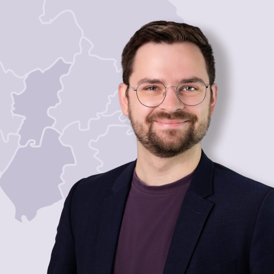 Robin Meier lächelt in die Kamera, trägt Brille, Bart und einen dunklen Blazer vor einer stilisierter Landkarte