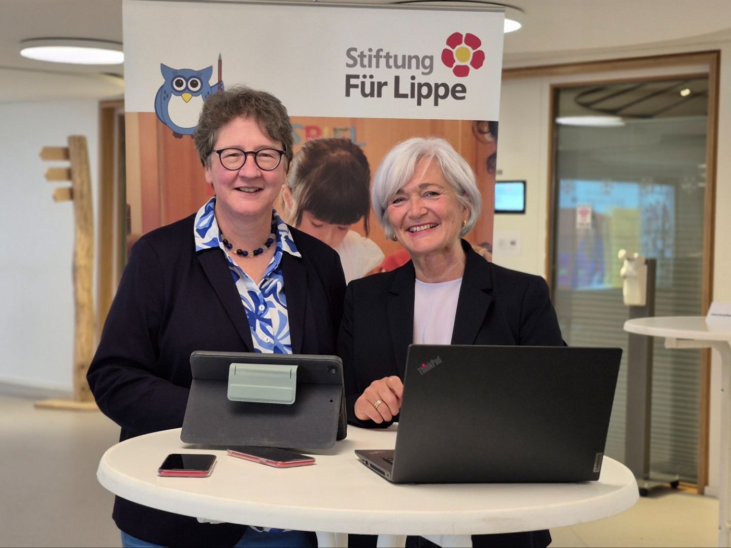 Zwei lächelnde Frauen stehen vor einem Banner der Stiftung Für Lippe mit digitalen Geräten auf einem Tisch