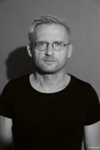 Ein Mann mit kurzen hellen Haaren, runder Brille und kurzem Bart trägt ein dunkles T-Shirt und blickt direkt nach vorn