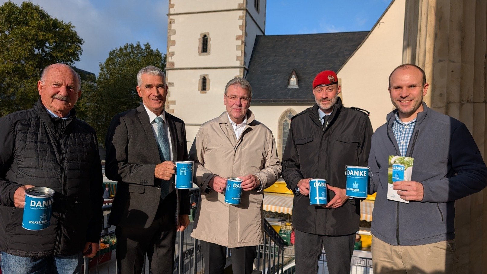 Fünf Männer präsentieren blaue Sammelbehälter und eine Broschüre des Volksbunds vor einer Kirche