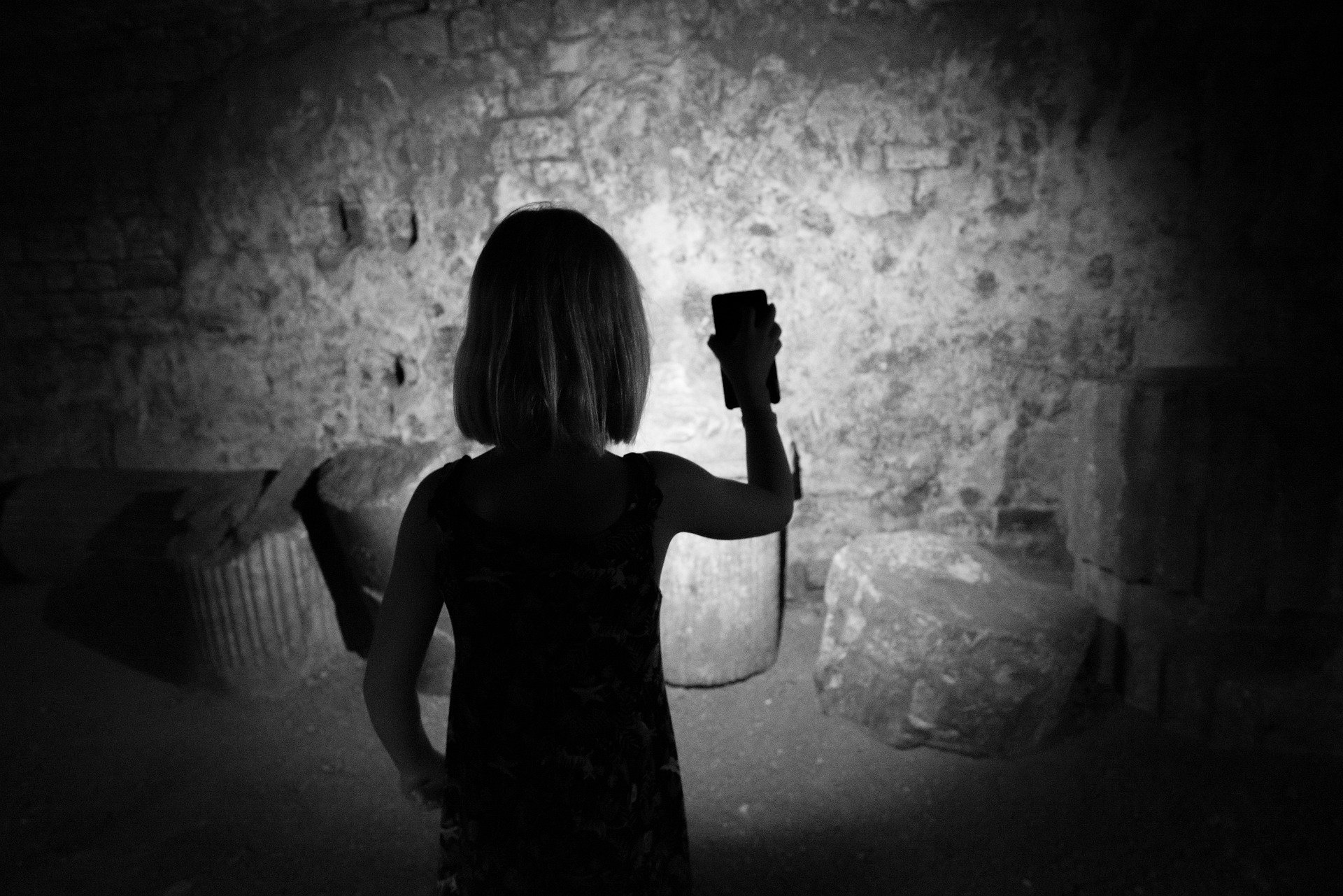 Person von hinten leuchtet mit Smartphone eine raue Steinmauer in einem dunklen Raum an
