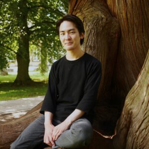 Teppei Kuroda sitzt auf den Wurzeln eines großen Baumes in einem Park, die Hände auf den Knien, und blickt in die Kamera