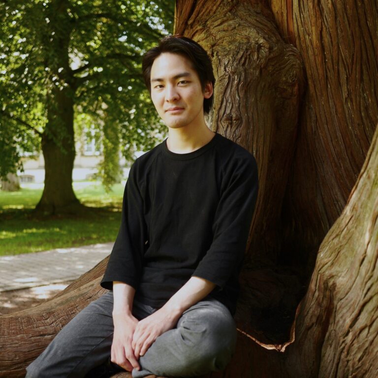 Teppei Kuroda sitzt auf den Wurzeln eines großen Baumes in einem Park, die Hände auf den Knien, und blickt in die Kamera