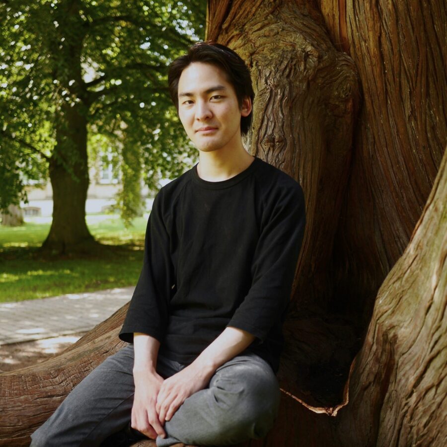 Teppei Kuroda sitzt auf den Wurzeln eines großen Baumes in einem Park, die Hände auf den Knien, und blickt in die Kamera