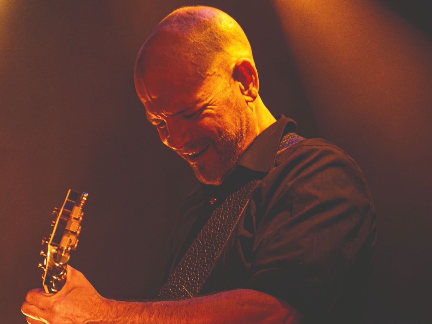 Tom Lüneburger mit Glatze und Bart lächelt und spielt Gitarre auf einer Bühne unter warmem Licht