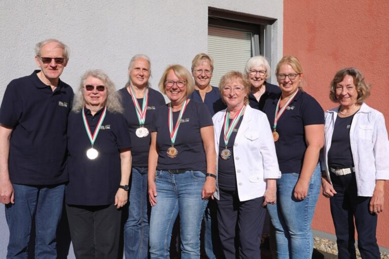 Neun lächelnde Personen in VIVa.OWL Poloshirts tragen Medaillen um den Hals und stehen vor einer Hauswand