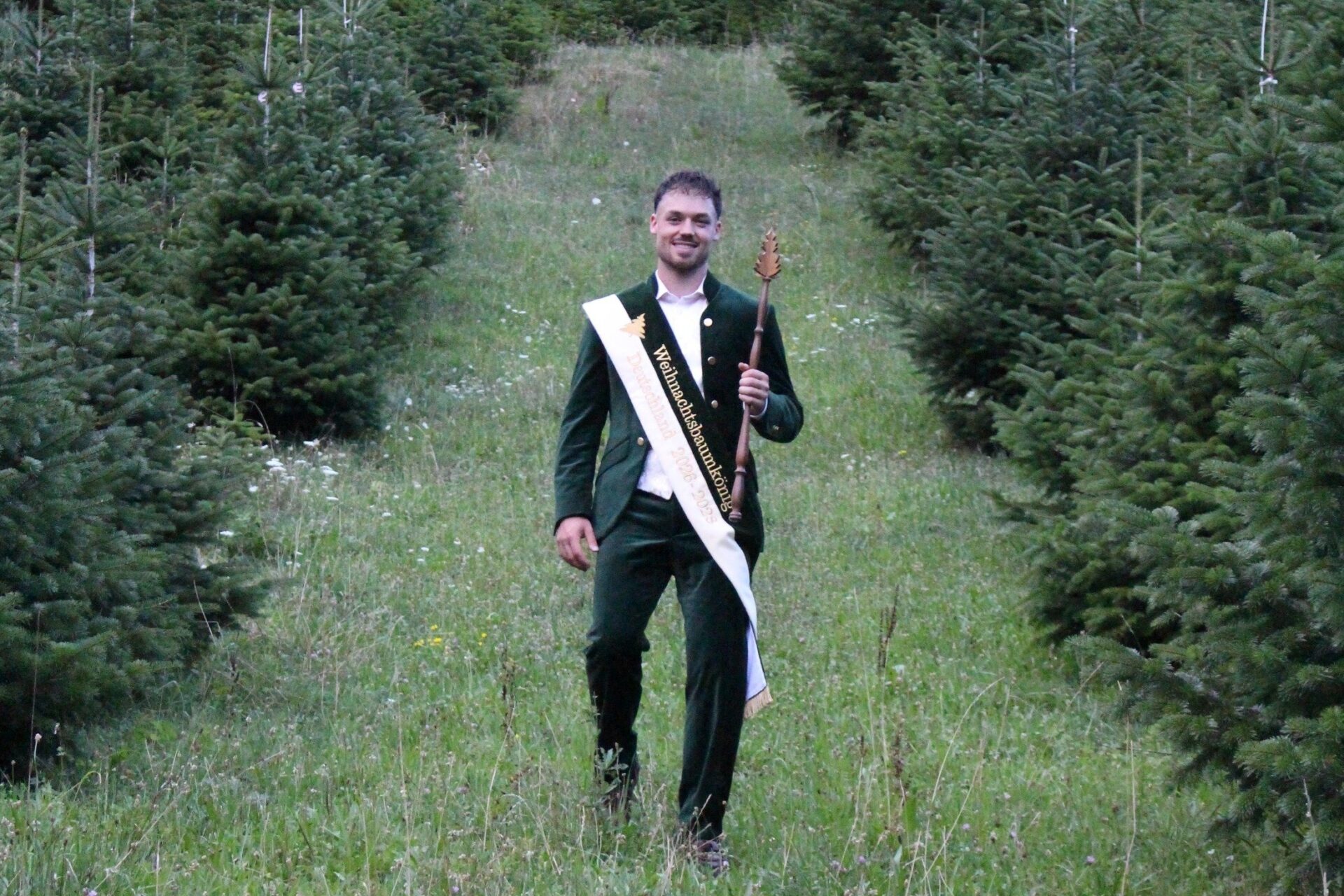 Der lächelnde Weihnachtsbaumkönig mit Schärpe und Zepter geht durch eine Wiese voller junger Tannenbäume