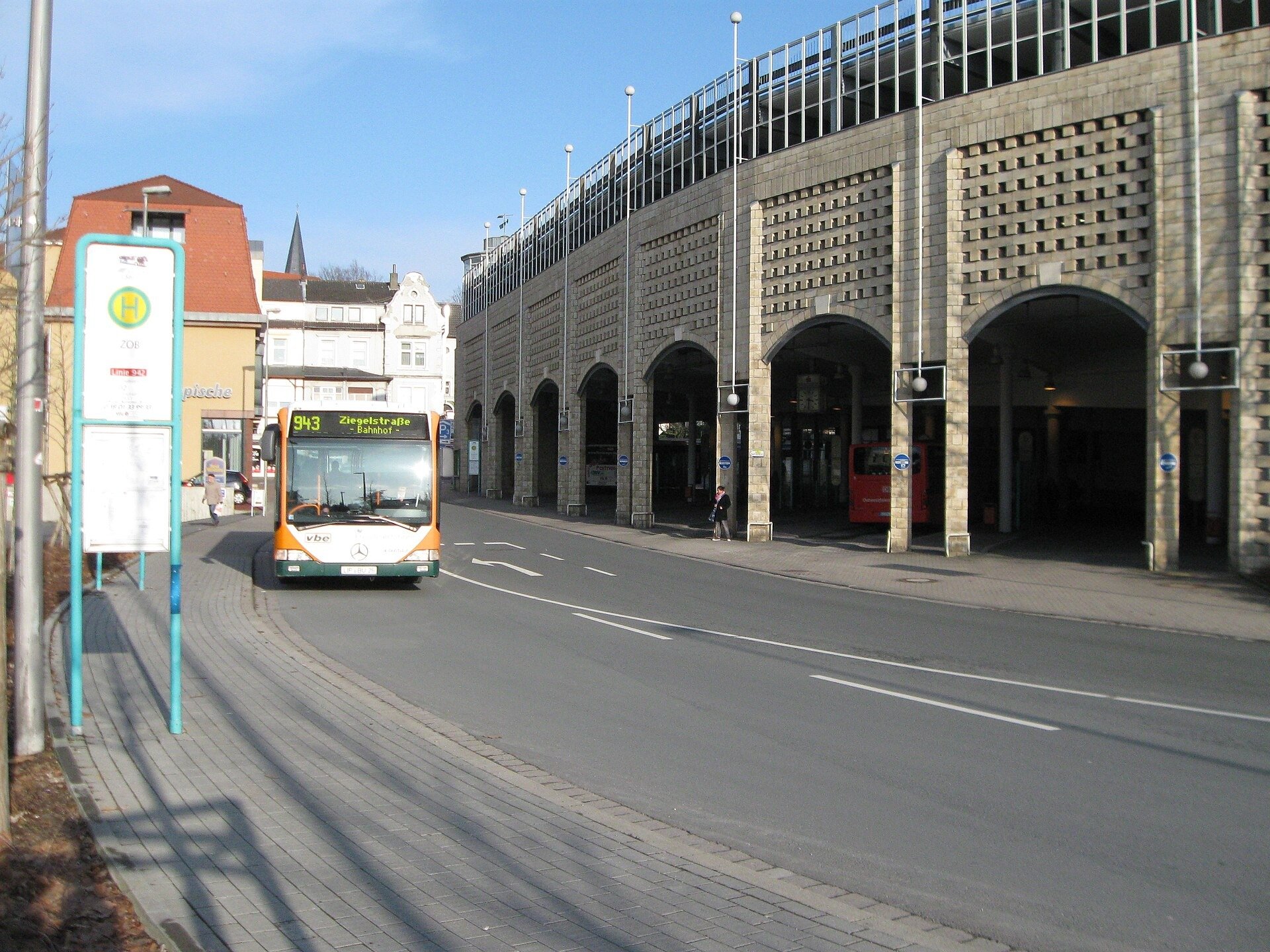 Ein orange-grüner Bus fährt auf einer Straße an einer Bushaltestelle und einem großen Steingebäude mit Bögen vorbei