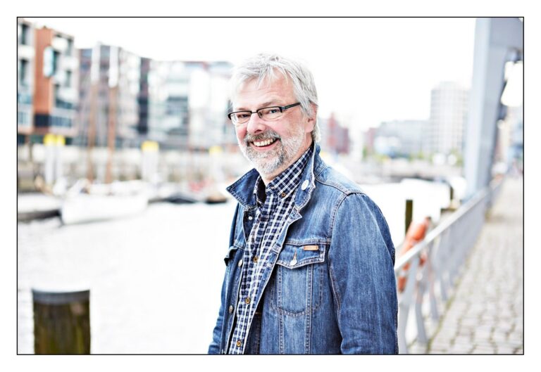 Der lächelnde Autor Alexander Häusser mit Bart und Brille in Jeansjacke am Wasser mit Gebäuden im Hintergrund