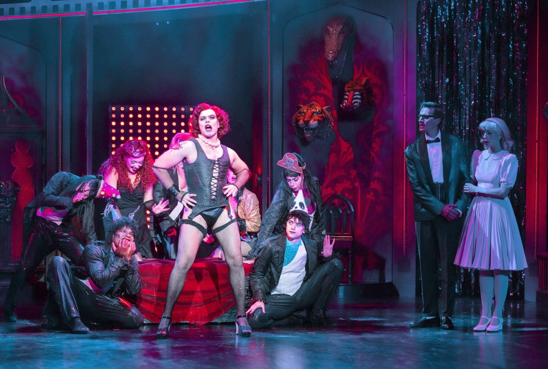 Frank-N-Furter in Korsett und Netzstrümpfen steht auf der Bühne, umgeben von schräg gekleideten Darstellern und Brad